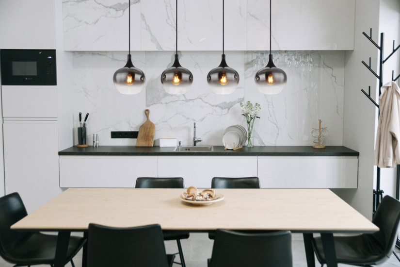 Moderne Küche mit heller Marmorwand, schwarzer Arbeitsplatte und fünf schwarzen Pendelleuchten über einem großen Esstisch aus Holz mit schwarzen Stühlen – stilvoll und minimalistisch eingerichtet